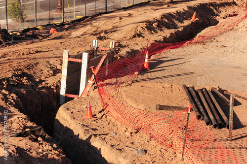 A dirt lot at a construction site, with an open ditch being excavated for a building foundation, showcasing uninstalled piping, an orange safety net, and cones to alert workers to the hazardous area photo