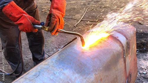 The welder cuts large metal pipes with ocetylene welding. A worker on the street cuts large-diameter pipes during the day and sparks and fire fly.