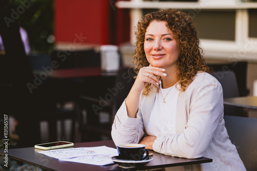 Woman sitting at an outdoor cafe. On the table are a smartphone, a coffee cup, and some papers