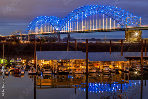 The photo showcases the illuminated Hernando de Soto Bridge spanning the Mississippi River, glowing in blue and white lights against a twilight sky. In the foreground, a marina with docked boats.