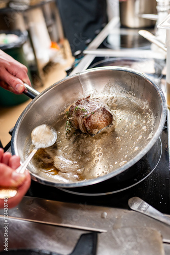 A chef's hands skillfully sear a piece of meat in a stainless steel pan, surrounded by sizzling butter and herbs. The kitchen atmosphere is dynamic and focused on culinary artistry.