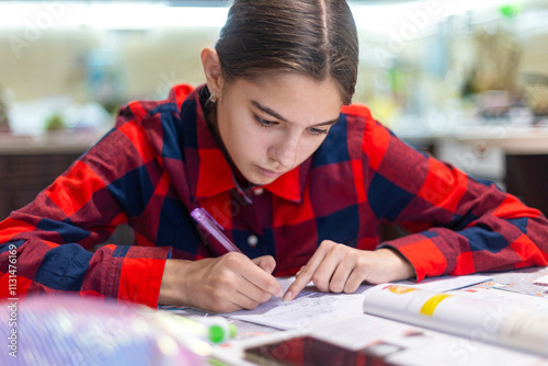 Focused student studying at home. A young girl wearing a red checkered shirt intensely studying at a desk with notebooks and a pen in a home setting.