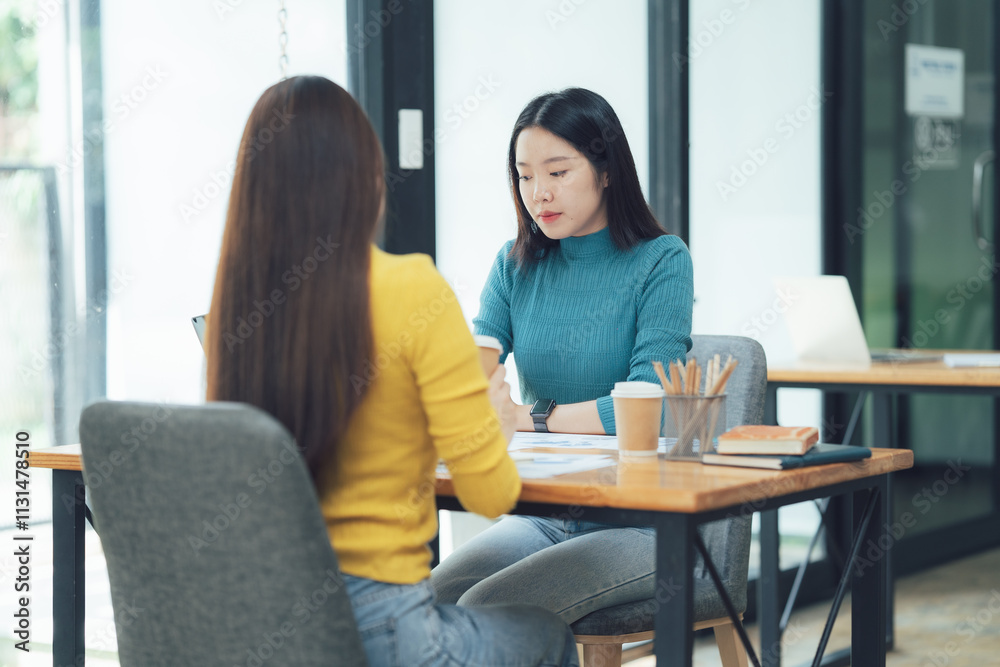 Fototapeta premium Collaborative Meeting: Two young Asian women engage in a focused discussion, leaning in attentively during a collaborative meeting at a modern office desk.