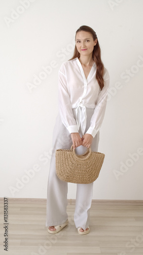 oung beautiful caucasian woman with long brunette hair, wearing white shirt, light grey pants, woven bag and sandals, shows her outfit. Women clothes.
