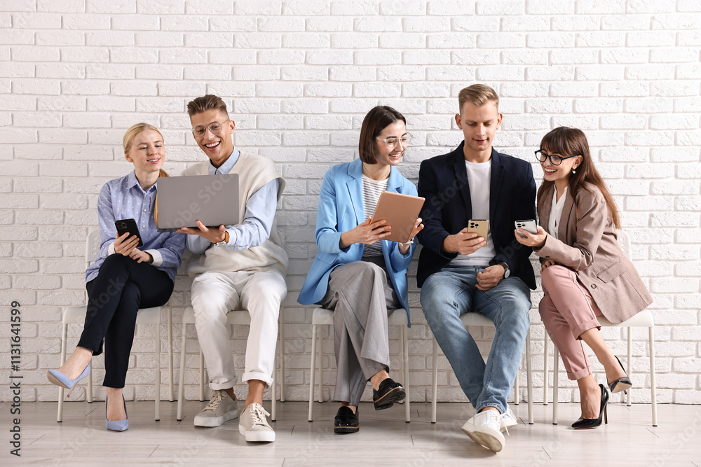 Fototapeta premium Group of people using different gadgets on chairs near white brick wall indoors. Modern technology