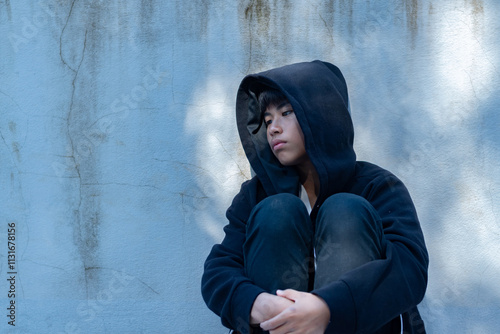 Asian pre-teen boy in a black hoodie sits with his knees pressed against a metal fence panel in a juvenile detention facility, awaiting further release, freedom and detention of people concept. photo