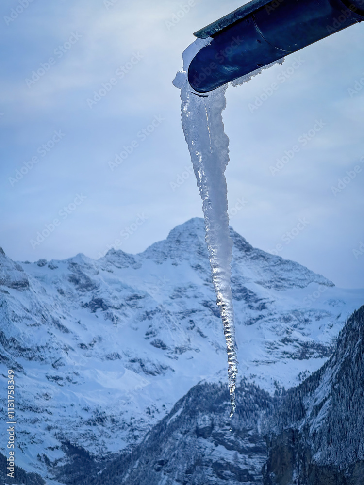 Icicle coming out of a gutter infront of snowcapped Breithorn mountain in Switzerland in winter against cloudy sky