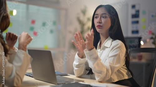 Two women are sitting at a desk with a laptop in front of them. One of the women is talking to the other