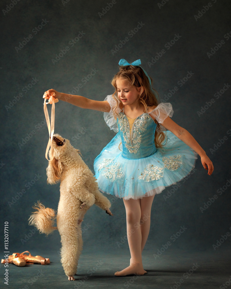 A ballerina girl in a ballet skirt and pointe shoes poses with poodle dog. Little adorable child ballerina in ballet tutu with puppy practicing classic choreography in ballet school, in studio