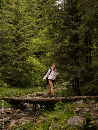 Young woman in a hoody and a packpack crossing bridge over the river in thick woods. Females adventure in the forest finding a river with a wooden bridge. Holidays activity inspiration. photo