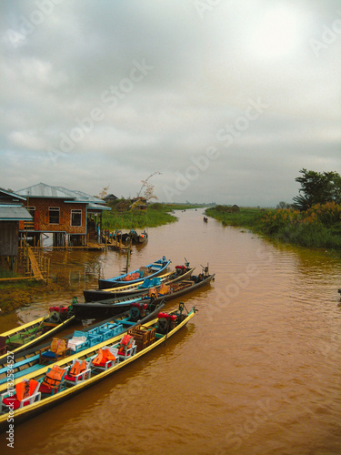 Wallpaper Mural  Inle Boat Station in Inle Nyaung Shwe Canal. A series of fishing boats along the river generated by Inle Lake. Homes and Pagoda temple in the background.Daily life in Inlay town, boat station to visi Torontodigital.ca