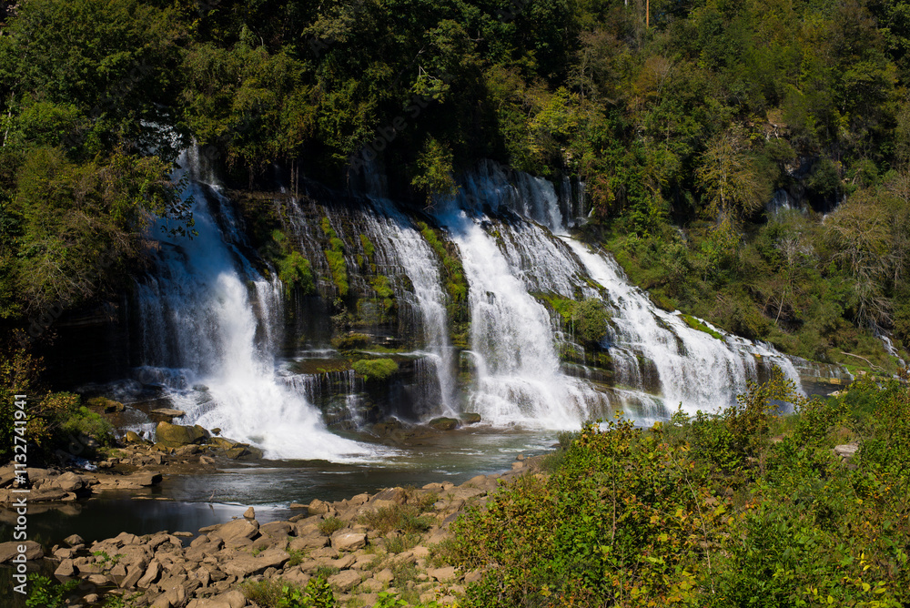 Fototapeta premium A stunning multi-tiered waterfall cascading down rocky cliffs surrounded by lush green foliage, captured on a sunny day.
