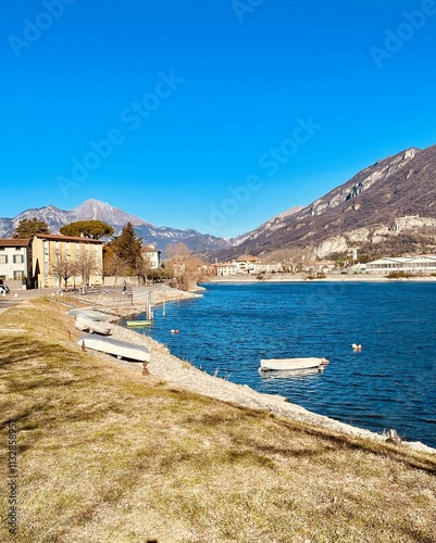 A scenic riverside view on a bright and sunny day, with vibrant blue water, small boats floating near the shore, and a backdrop of picturesque mountains. A row of houses and buildings on the likeside photo