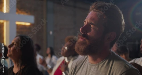 Bearded man praying during church worship, hands raised in devotion, illuminated cross and diverse congregation in background, expression of faith and spirituality
