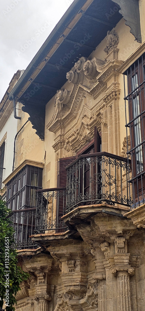 Fototapeta premium Historical building facade with ornate baroque details, iron balconies, and decorative columns; worn plaster and wooden shutters complement the architectural style;