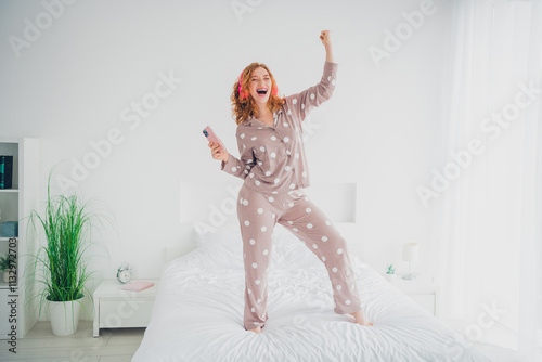 Joyful young woman with red hair dancing in cozy bedroom wearing polka dot sleepwear, celebrating morning energy with smartphone in hand photo