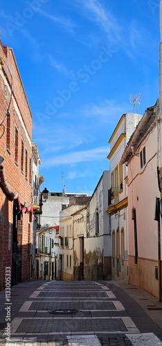 A narrow cobblestone street with historic buildings on both sides; red brick on the left and white facades with yellow details on the right under a clear blue sky;