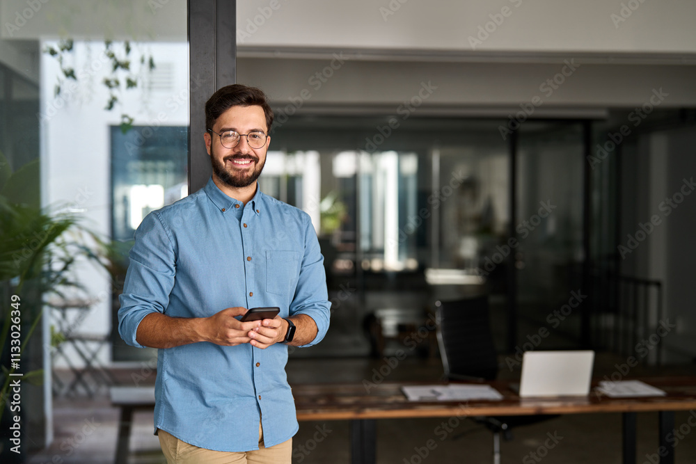 Obraz premium Happy professional Latin young business man looking at camera at work, portrait. Confident smiling businessman leader, male executive manager entrepreneur standing in office with smartphone in hands.
