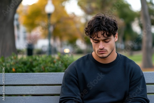 A young man sits pensively on a bench in a park, looking downwards with a thoughtful expression, capturing a moment of quiet reflection amid nature's beauty.