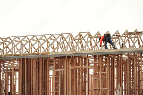 A roof carpenter atop a two-story wooden home under construction leans over to secure plywood sheets to the roof structure with a pneumatic framing nailer photo