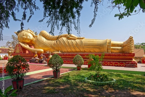 The reclining Buddha statue at Phi That Luang temple in Vientiane, Laos, which is surrounded by potted plants and a brick walkway.
