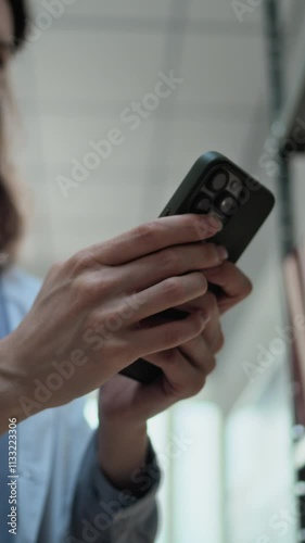 Vertical shot of female scientist in lab coat chatting, surfing the Internet or social media on mobile phone while standing near shelf with folders. Modern archaeological science laboratory. Low Angle photo