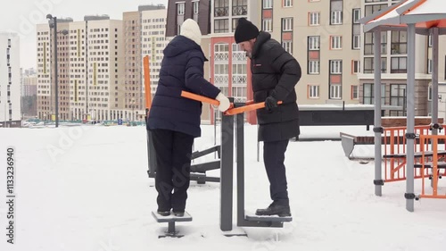 Elderly couple in warm clothing exercising on outdoor equipment in a snowy urban park. Healthy lifestyle and active retirement life concept