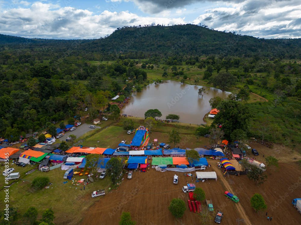 Fototapeta premium Aerial View of Rural Market by the Lake: A picturesque aerial shot captures a vibrant rural market nestled beside a serene lake, surrounded by lush green hills under a partly cloudy sky.