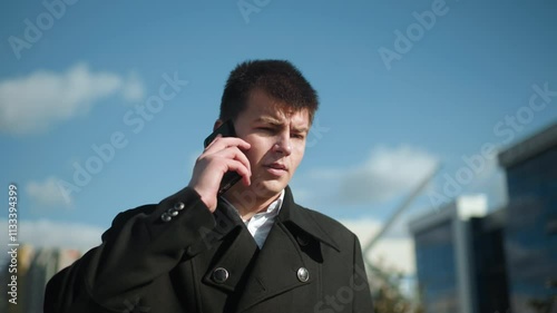 Businessman wearing black coat with metallic buttons, on phone call outdoors under bright sky, looking contemplatively with blurred modern buildings and greenery in background photo