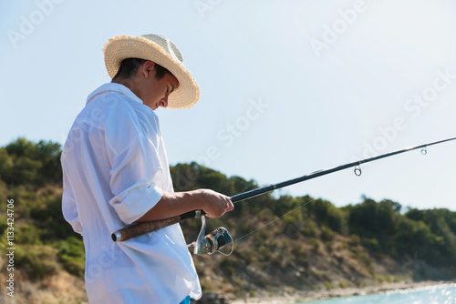 A teenage boy in a straw hat and white shirt is holding a fishing rod while standing near the sea with lush greenery in the background under a clear blue sky photo