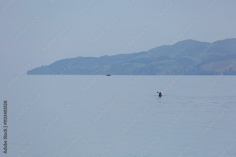 Obraz premium Kayaker paddles across calm water, serene landscape. Peaceful morning on the lake. NGARIMU BAY, THAMES, COROMANDEL PENINSULA, NEW ZEALAND