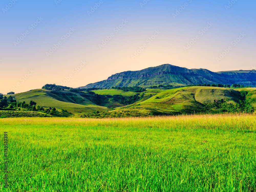Drakensberg mountain peak against a warm sunset sky, with a green valley grassland in the foreground, KwaZulu-Natal, South Africa