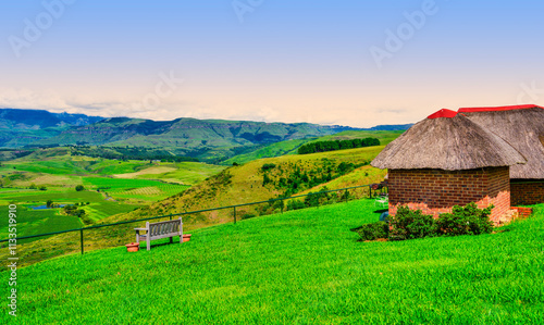 Obraz na płótnie Hilltop house with a thatched roof and a bench on the edge, overlooking the Drakensberg Mountain and the valley below, KwaZulu-Natal, South Africa w salonie