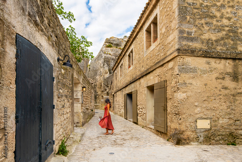 Fototapeta Naklejka Na Ścianę i Meble -  A barefoot young woman in a red dress walks along a cobblestone street with her small dog in the idyllic medieval and ancient town of Les Baux-de-Provence, France.