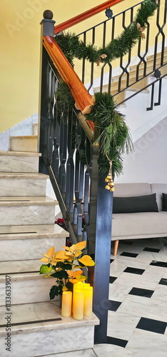 Staircase decorated with pine garlands and small golden bells; yellow poinsettias and three LED candles placed at the base; tiled floor with black and white pattern;