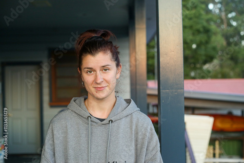 A casual portrait of a young woman wearing a grey hoodie, standing on a porch with a relaxed expression. The background features a mix of indoor and outdoor elements, including greenery and buildings