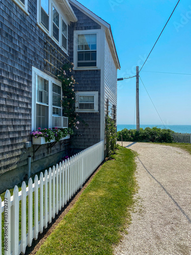 A coastal home with shingle siding, a white picket fence, blooming flower boxes, and a path leading to the ocean under a clear blue sky, embodying serene seaside charm.