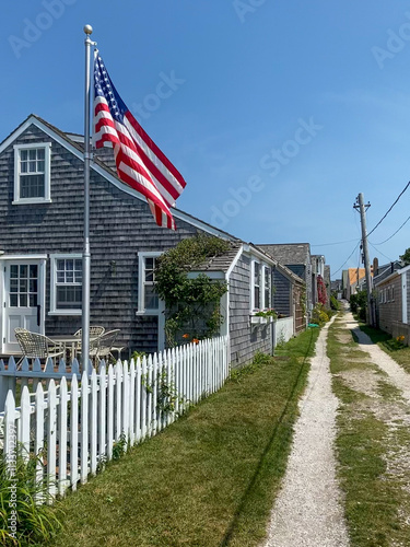 A quaint coastal cottage with shingle siding, an American flag, white picket fence, and a narrow gravel path, capturing a serene and patriotic seaside atmosphere under a clear blue sky.