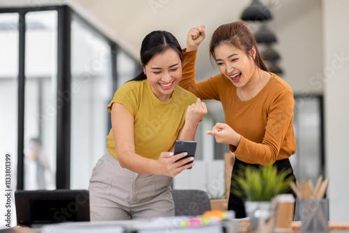 Two asian young women use laptop to work at workplace and lift her arm showing happiness teamwork celebrating achievement and success concept. photo