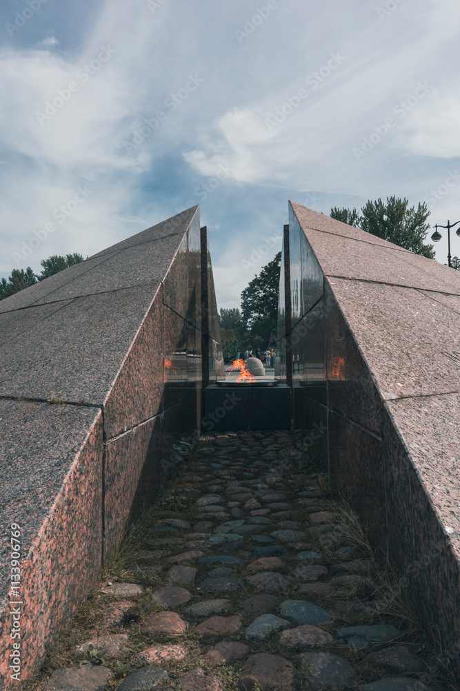 Obraz premium A solemn monument featuring an eternal flame between granite walls, with a cobblestone path leading through, symbolizing remembrance and respect against an open sky