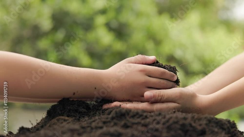 in a gentle act of love, the younger sibling pours dark fertile soil into the waiting hands of the elder, reflecting hope, care and the promise of nurturing the earth together, green garden background