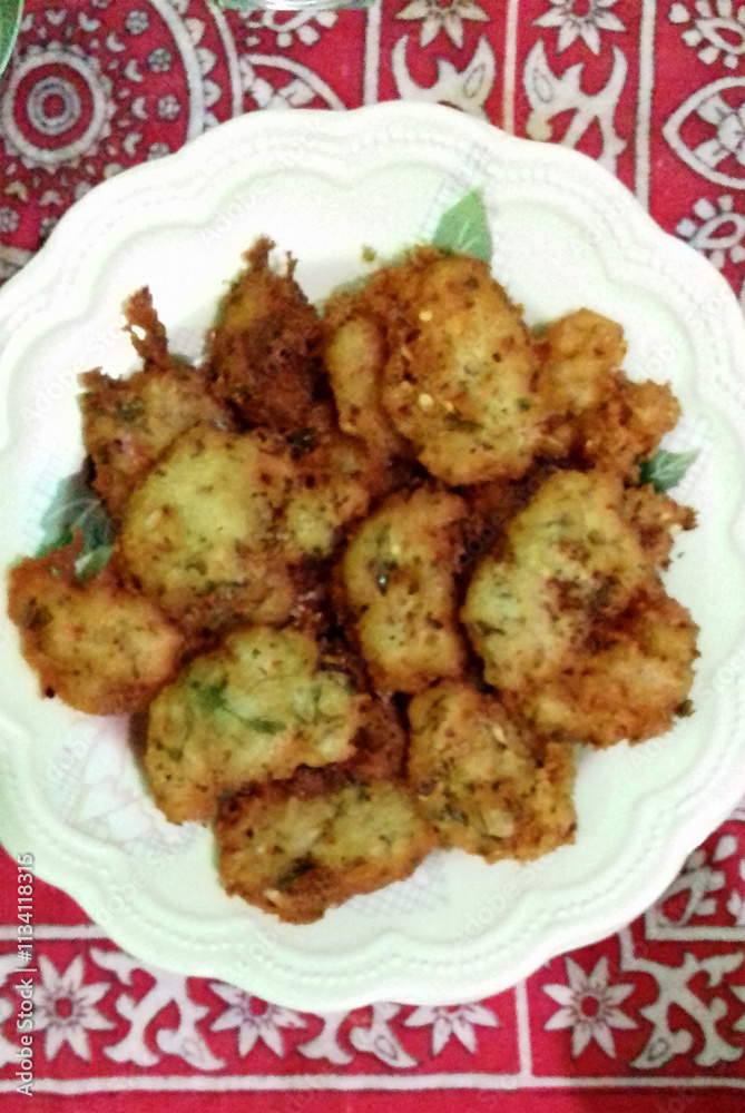 Golden-brown fritters are piled on a white plate with decorative green accents, sitting on a red patterned tablecloth. The crisp edges and light seasoning suggest a flavorful snack. Golden-brown fritters are piled on a white plate with decorative green accents, sitting on a red patterned tablecloth. The crisp edges and light seasoning suggest a flavorful snack.
