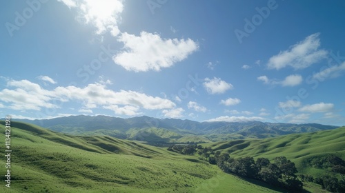 Scenic valley landscape with rolling hills, lush green grass, and a vibrant blue sky dotted with fluffy clouds.