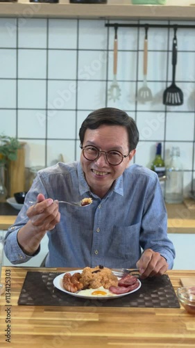 Happy middle aged man eating fried rice with sunny side up egg and sausages in warm kitchen