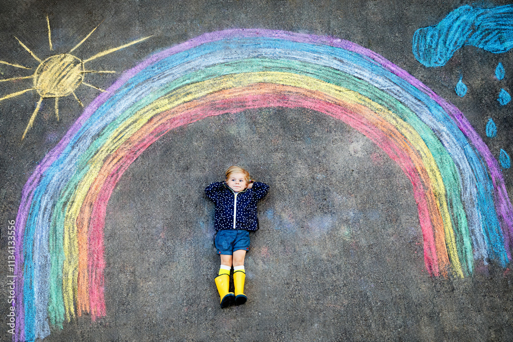 Happy little toddler girl in rubber boots with rainbow sun and clouds with rain painted with colorful chalks on ground or asphalt in summer. Cute child having fun. creative leisure