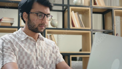 A young bearded Indian south Asian corporate office man or male employee or executive in formal wear and eyeglasses is busy typing and working on a laptop in a modern start up business workplace. 
