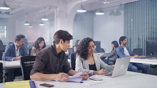 Two young modern male and female corporate employees discussing and working in a team together on an assignment using a laptop with office colleagues sitting in a busy start up business workplace.