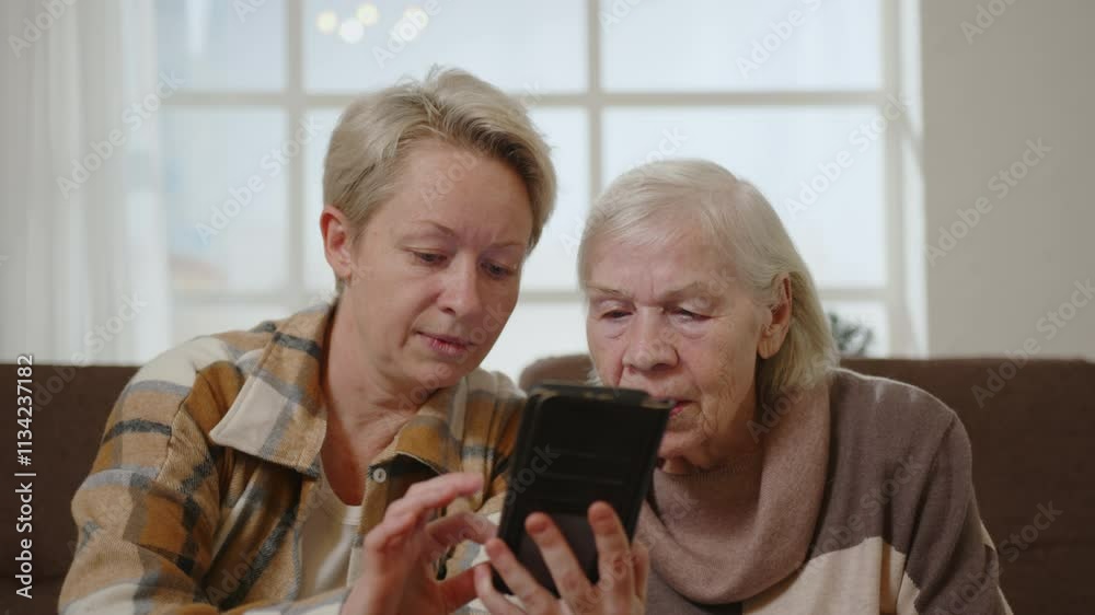 Two women elderly mother and middle aged daughter using smartphone while sitting on sofa in room. Two women elderly mother and middle aged daughter using smartphone while sitting on sofa in room.