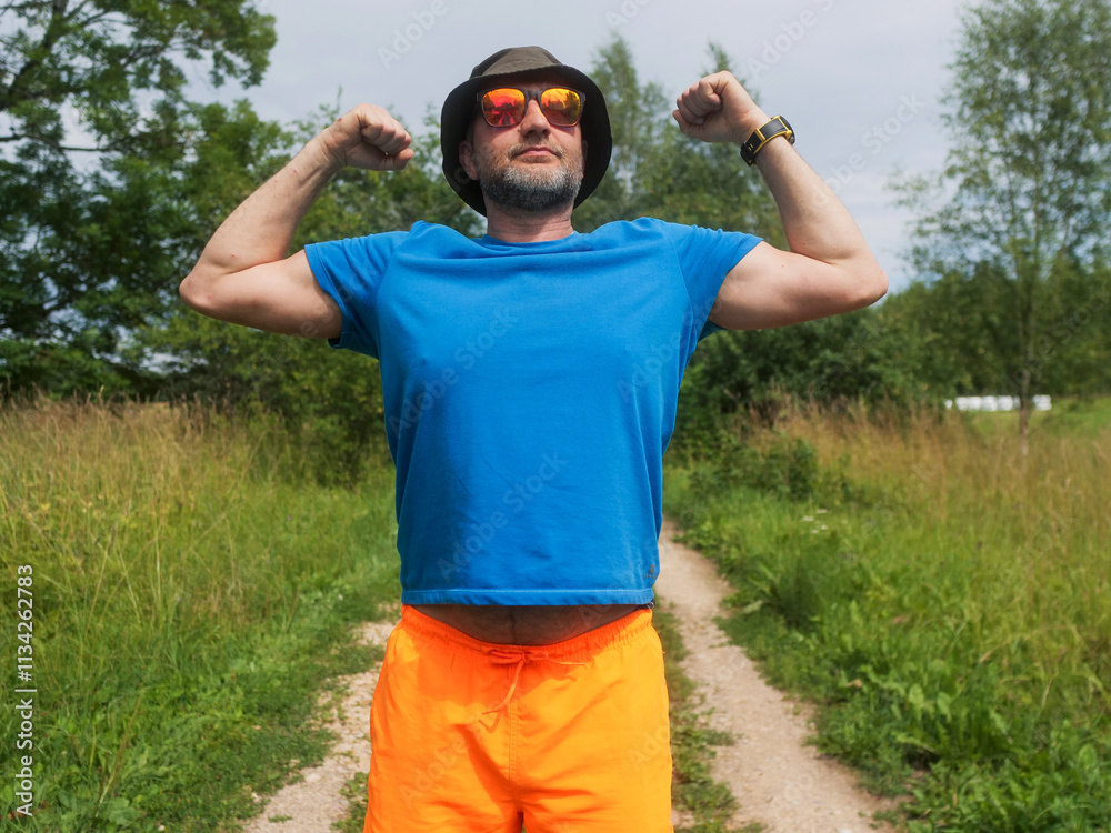 Naklejka premium A man in a blue shirt and orange shorts flexes his hands. The man is wearing a hat and he is enjoying the outdoors. Male in his 40s with grey beard. Sport and fitness outdoor.