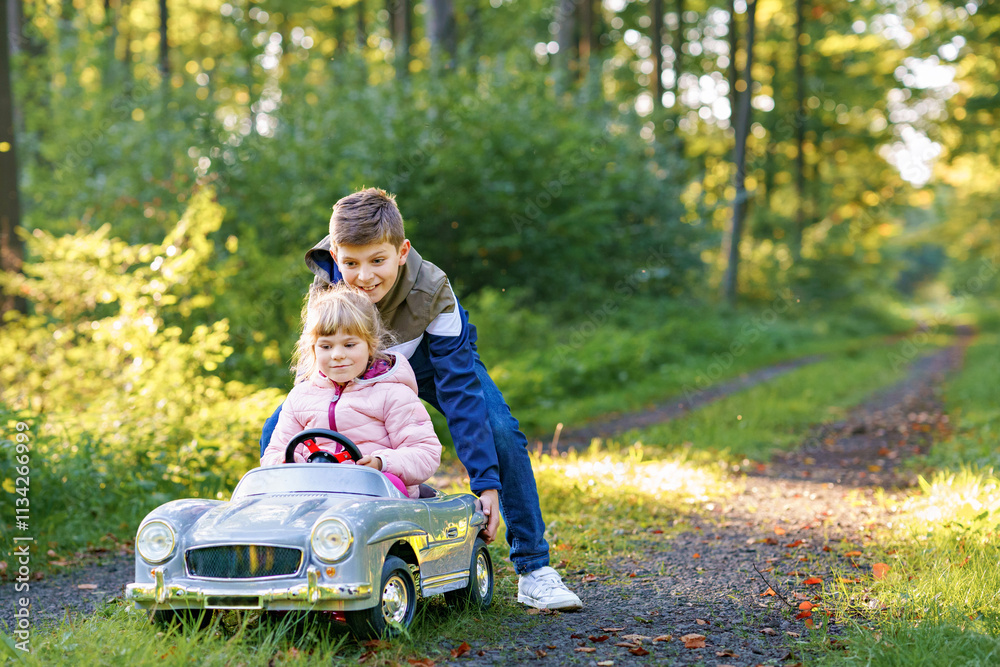 Two happy children playing with big old toy car in autumn forest, outdoors. Kid boy pushing and driving car with little toddler girl, cute sister inside. Laughing and smiling kids. Lovely family